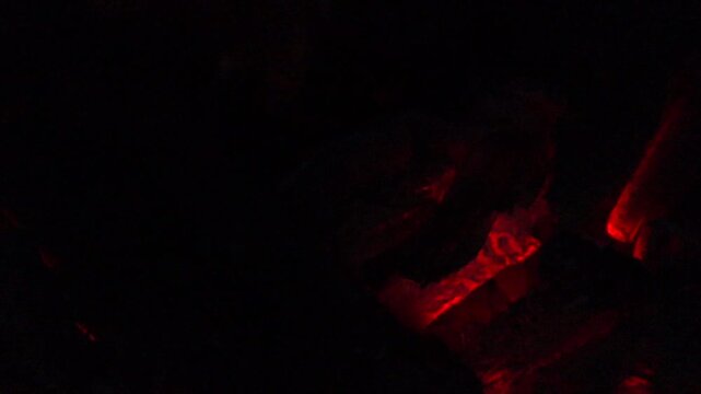 Close-up of glowing red embers and a small flame inside a dark fire pit, creating a dramatic scene of heat, combustion, and burning wood textures.