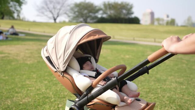 Baby sleeps calmly in stroller. Adult gently pushes stroller through green park. Sunlight glows softly on grassy field. Baby's peaceful face shows contentment. Perfect for parenting, family