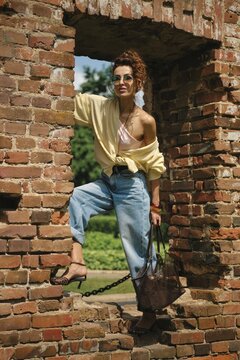 A stylish woman stands with one foot resting on a brick wall, dressed in casual fashion