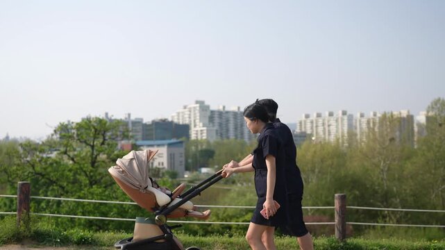 Two women push a stroller along a park path. They walk side by side, smiling at each other. City buildings rise behind green trees and grass. Sunlight glows over the tranquil stroll