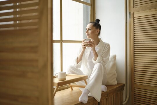 Dreamy woman in white clothes sitting on windowsill and holding cup of coffee or tea with both hands