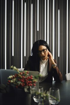 A businesswoman is seated at a desk in a contemporary office, deep in thought, with stylish vertical lighting casting soft shadows. She is dressed in professional attire and appears focused