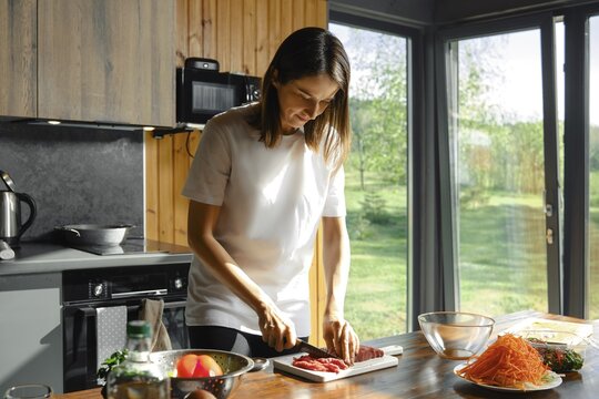 Cute woman is slicing fresh beef meat on a cutting board in a modern kitchen filled with natural light. The outdoor view complements her healthy meal preparation