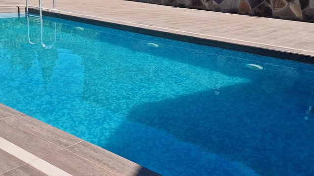 Rectangular outdoor swimming pool with crystal clear blue water, mosaic tile bottom, and a modern wooden texture tiled deck on a sunny day. Luxury backyard or resort area