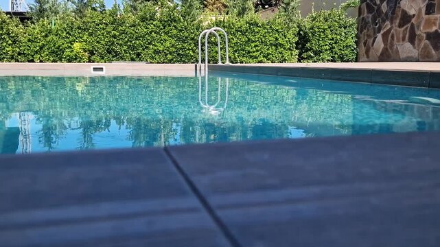 Rectangular outdoor swimming pool with crystal clear blue water, mosaic tile bottom, and a modern wooden texture tiled deck on a sunny day. Luxury backyard or resort area