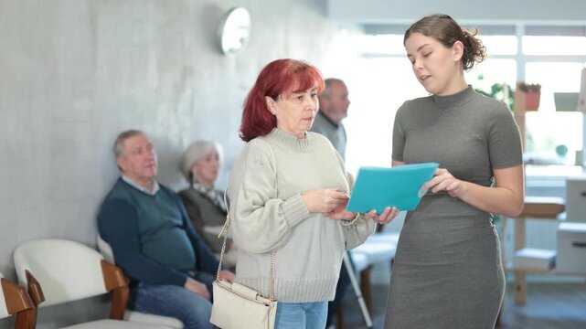 Positive polite young secretary or social worker talking to aged woman visitor in reception area in background queue of people