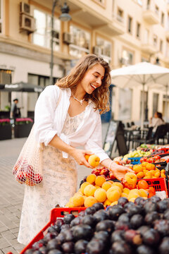 A smiling woman in a dress chooses fresh fruit at a market stall. A beautiful woman puts fresh fruits and vegetables into a string bag at a fruiteria. Concepts: food, shopping, weekend.