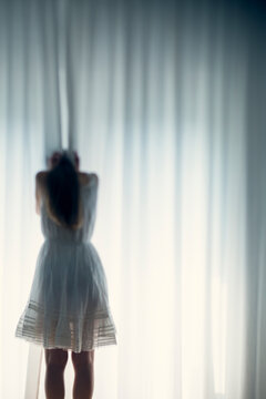 Young woman hiding her face behind a white curtain, back view