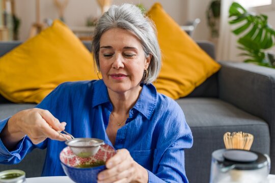Woman preparing matcha tea with a strainer and whisk in the living room