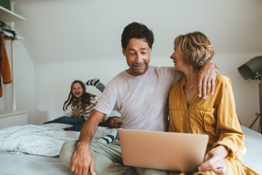 Man with laptop sitting by woman looking at daughter and son playing on bed at home