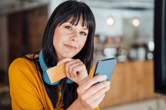 Smiling woman with with headphones around neck holding mobile phone and credit card in cafe