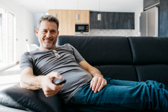 Smiling man holding TV remote while sitting on sofa at living room