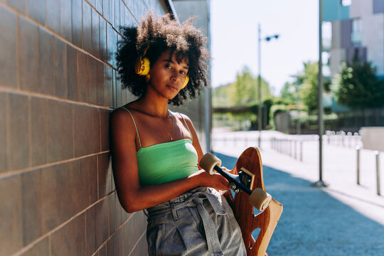 Young woman wearing headphones holding skateboard leaning on wall