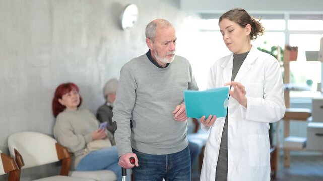 Positive polite young nurse or doctor talking to aged man patient in reception area in background queue of people