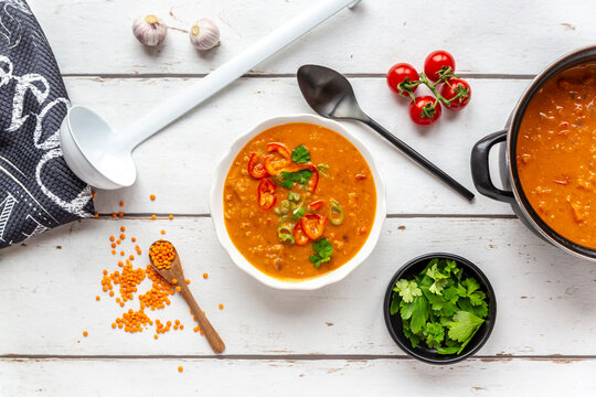 Bowl and pot of garnished red lentil soup and ingredients on white wood