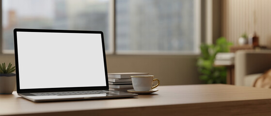 Fototapeta premium White screen laptop with books and coffee cup on wooden table across glass window in the living room