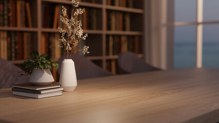 Wooden table with books, flower vase and potted plant across bookshelf and window in dim lit library © bongkarn