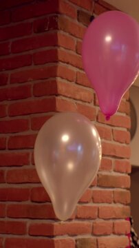 A pink and a pearlescent helium balloon float near the center-right of an indoor frame next to a textured red brick column. Soft warm lighting creates gentle