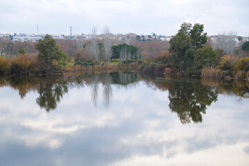 Obraz premium Scenic view of the Guadiana River with perfectly calm water creating symmetrical tree and cloud reflections next to the Guadiana River Park in Merida, Extremadura, Spain.