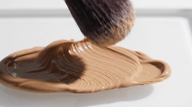 Close up of professional makeup brush applying liquid foundation and concealer cream on a white surface for cosmetic advertising