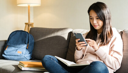 Obraz premium Tapping student wearing hoodie, jeans on sofa at home with smartphone and open book, copy space