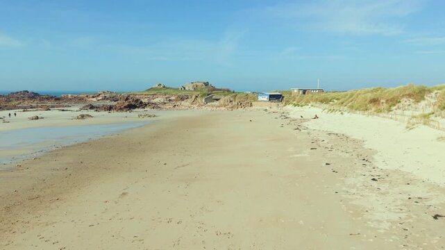 La Grosse Rocque beach Cobo Bay Guernsey.Low drone flight along beautiful sandy beach and sand dunes at low tide on shoreline towards rocky granite headland with military fort in late afternoon sun