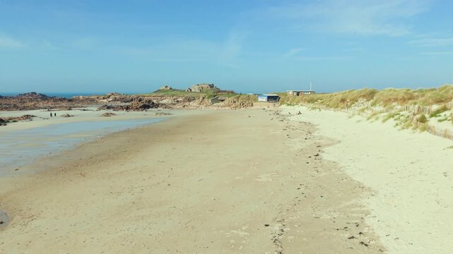 La Grosse Rocque beach Cobo Bay Guernsey.Low flight along beautiful empty sandy beach and sand dunes at low tide on shoreline towards rocky granite headland with old military fort in afternoon sun