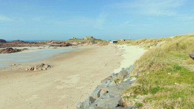 La Grosse Rocque beach Cobo Bay Guernsey.Low drone flight across beautiful sandy beach and sand dunes at low tide on edge of sea towards rocky granite headland with old military fort in afternoon sun