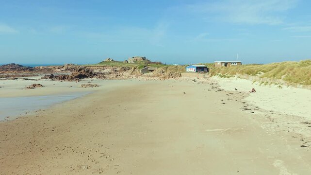 La Grosse Rocque beach Cobo Bay Guernsey.Low forward flight over quiet sandy beach and sand dunes at low tide on shoreline towards rocky granite headland with old military fort in late afternoon sun