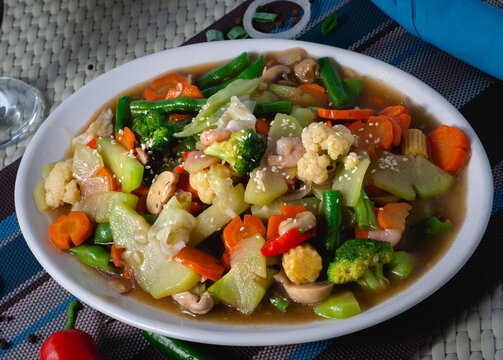 over head photo of chopsuey, a stir-fry of mixed vegetable pieces. Broccoli, carrots, red bell pepper, chayote, green beans, cauliflower, mushroom, young corn, meat and seafood bits on white platter. 