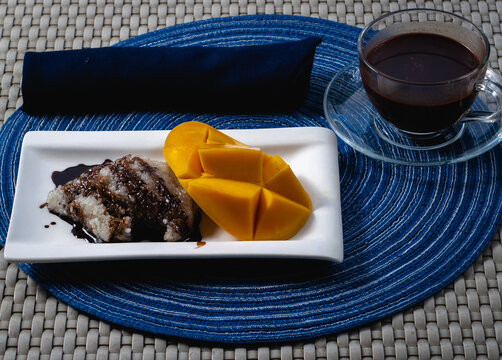 close up photo of puto maya and sikwati also known as sweet sticky rice with a glass of hot chocolate and a slice of ripe mango on a small rectangular white plate.
