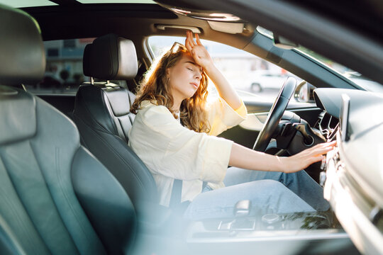 A young woman sits behind the wheel of a hot car. A female driver feels unwell due to the heat in the car on a sunny day. Concept of air conditioning, cooling, and transportation.