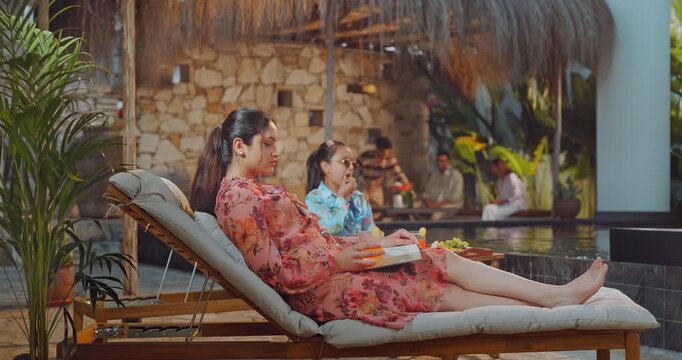 Indian young adult relax woman sitting on sun lounger chair read new novel book enjoy trip day female hold glass drink juice do self care girl child eat fruits spend time have fun joy pool edge place