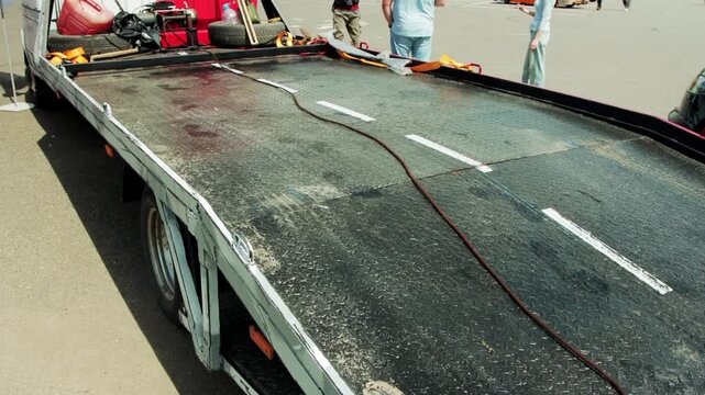 Close-up view of empty flatbed of car transporter truck, showing rugged diamond plate metal surface. White dashed lines imitate road markings. Dark power cable lies across platform. Background shows b