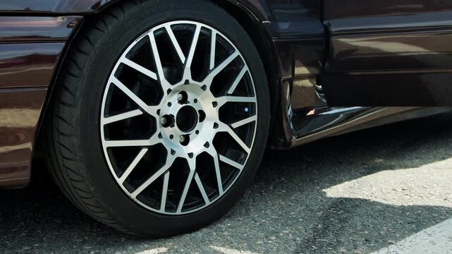 Detailed shot of shiny two-tone spoked rim with radial tire on dark metallic vehicle exterior, parked on textured gray pavement under natural daylight, highlighting automotive engineering detail