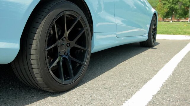 Exterior detail shot of light blue sports car or sedan showing front passenger side wheel. Black multi-spoke alloy rim and high-performance tire visible. Asphalt ground with white parking stripe in fo
