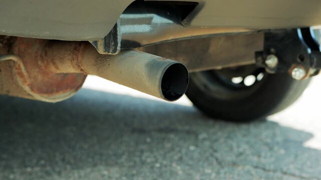 Close up view of car exhaust system, featuring tailpipe and rusty muffler. Image highlights vehicle undercarriage with tire background, detail of mechanical components on asphalt.