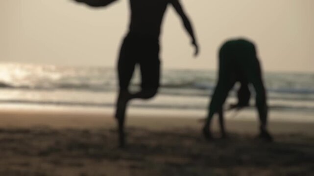 Silhouettes of two fighters practicing the Afro-Brazilian martial art of capoeira, with its fluid acrobatic movements and kicks, on a sandy beach against the warm light of the setting sun