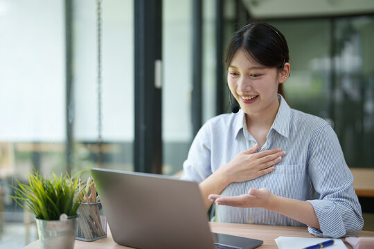 Young Asian woman wearing a headset and smiling while interacting with a laptop during a virtual meeting, actively communicating and gesturing, working remotely in a modern office setup