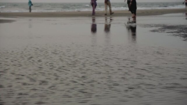 Serene view of anonymous people walking along the wet shoreline, with their reflections mirrored on the calm, shallow water as gentle waves roll in during an overcast day