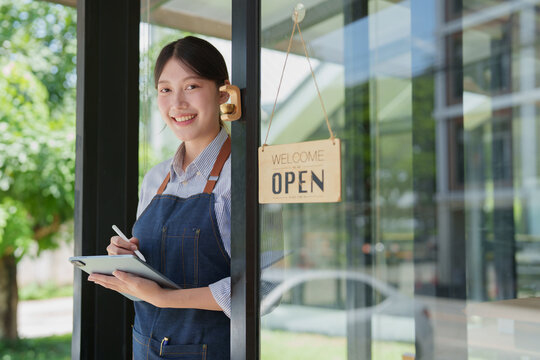 Smiling young asian woman shop owner holding a digital tablet with a stylus, standing at her cafe entrance with an open sign, ready to welcome customers