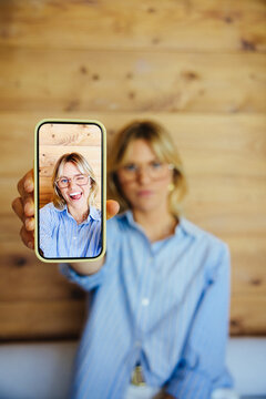 Woman holding smartphone displaying identical photo of herself on wooden background
