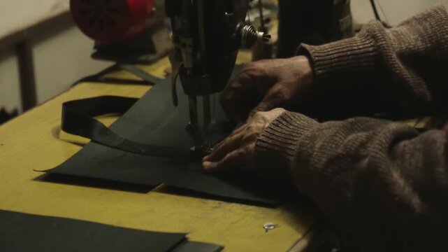 Skilled artisan's hands carefully guiding black fabric through a vintage sewing machine, meticulously stitching in a traditional workshop atmosphere illuminated by a warm single light source
