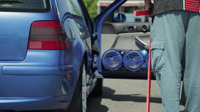 Close up view of blue modified car with open rear door showing multiple blue speakers person holding measuring device near vehicle for sound system evaluation during contest.