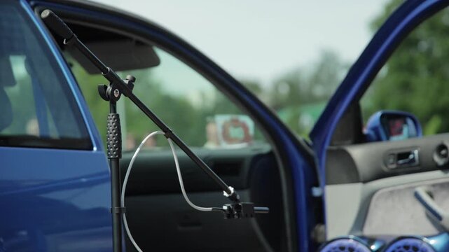Detail view of microphone and stand placed outside open door of vibrant blue car, ready for car audio sound quality and decibel level assessment at outdoor event, highlights professional equipment for