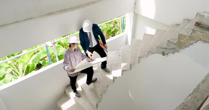 Overhead stairwell view presents Asian woman engineer and male contractor coordinating blueprint review, construction safety inspection, and building project progress tracking during site visit.