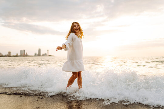 A woman strolls along the seashore at sunset. A beautiful woman in a white dress enjoys the seascape, feeling the freedom of the outdoors. Concepts of beauty, peace, and relaxation.