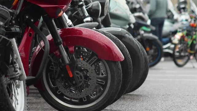 Close-up view of multiple motorcycles parked outdoors in a row, foreground highlighting front wheel, fender, and disc brake of a vibrant red cruiser motorcycle, rain droplets visible.