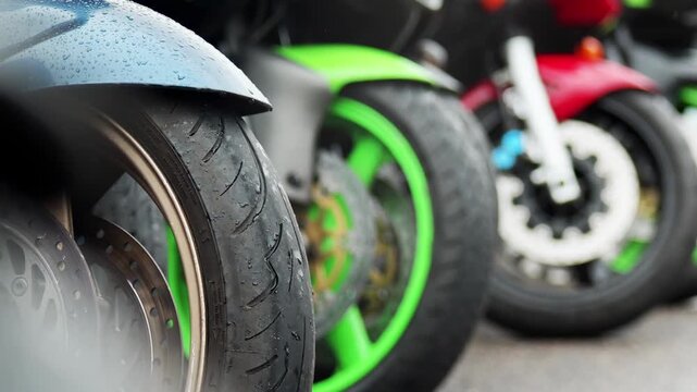 Focus on front section of blue, green, and red motorcycles side by side. Water droplets visible on fender of blue bike. Disc brakes and tire treads are prominent. Blurred background suggests outdoor s