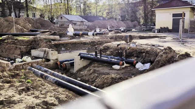 Extensive excavation site revealing complex network of black insulated pipes laid in deep trench, surrounded by mounds of soil and construction debris. Bright sunlight illuminates urban street scene w
